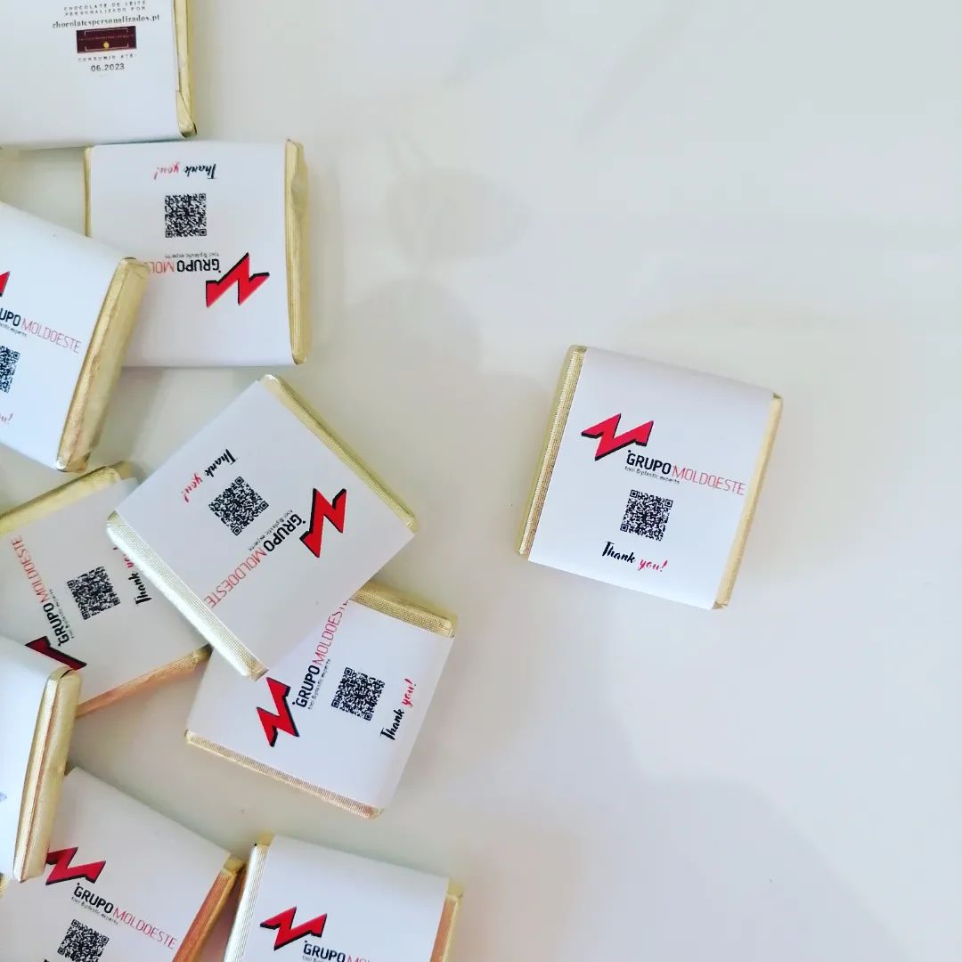 Pequenos chocolates com papel branco, etiqueta com logotipo vermelho, QR code e texto sobre fundo branco.