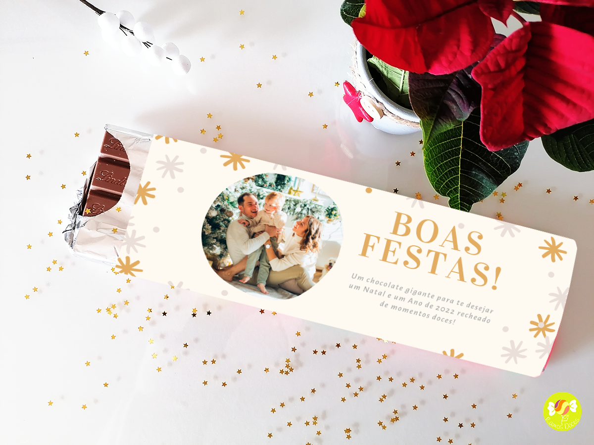 Barra de chocolate com embalagem branca, mensagem Boas Festas e foto de casal, decorada com estrelas douradas, ao lado de planta com flores vermelhas