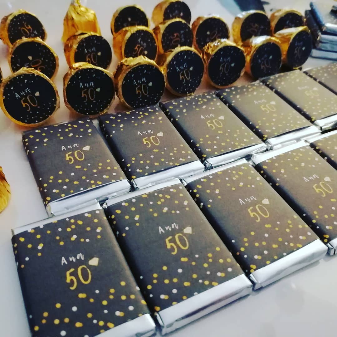 Chocolates embalados em papel preto com pontos dourados e texto Ana 50 e chocolates em papel dourado com rótulo preto com texto Ana 50