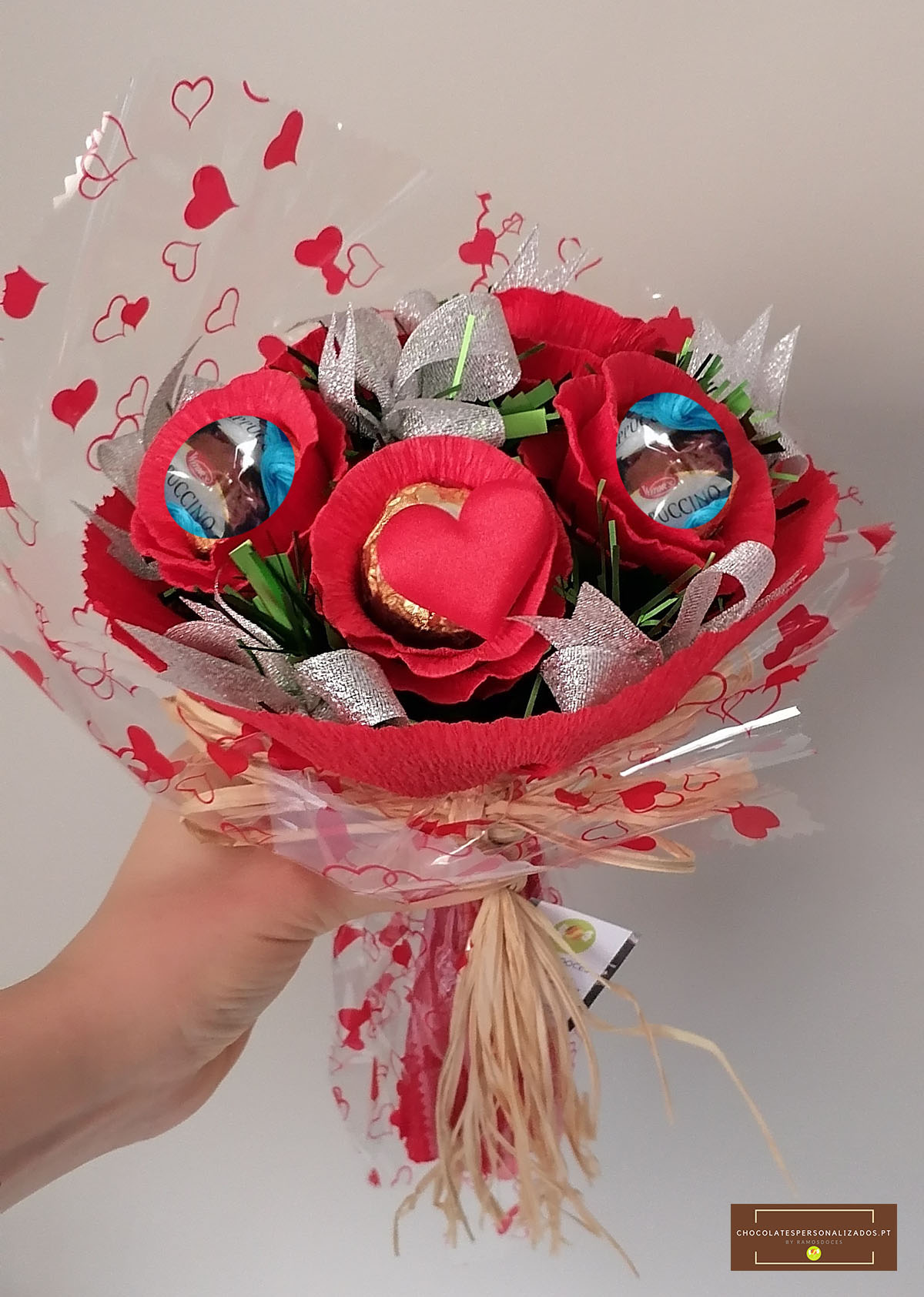 Buquê de flores vermelhas de papel com chocolates em embalagem azul e detalhes prateados, envolto em plástico com corações vermelhos.