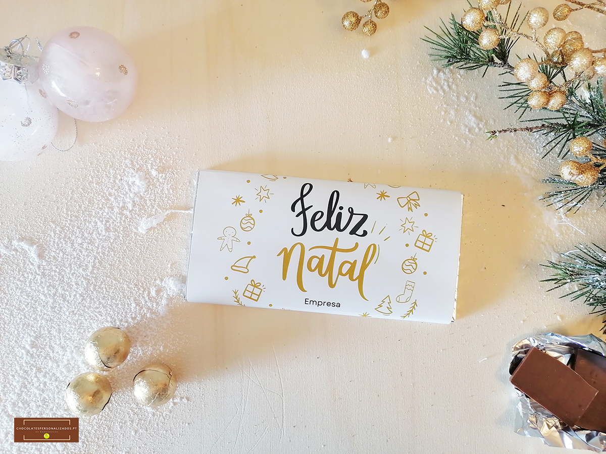 Barra de chocolate embrulhada com tema natalício em papel branco com texto Feliz Natal e decoração dourada ao redor