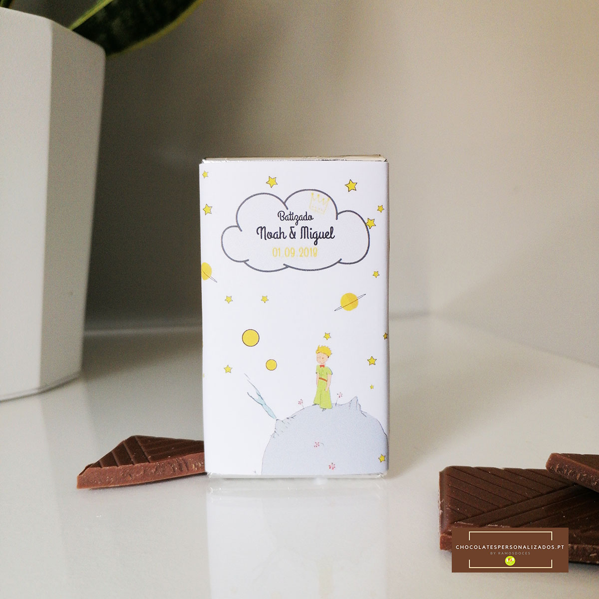 Embalagem branca de chocolate com ilustração do Pequeno Príncipe e texto de celebração, com pedaços de chocolate ao leite.