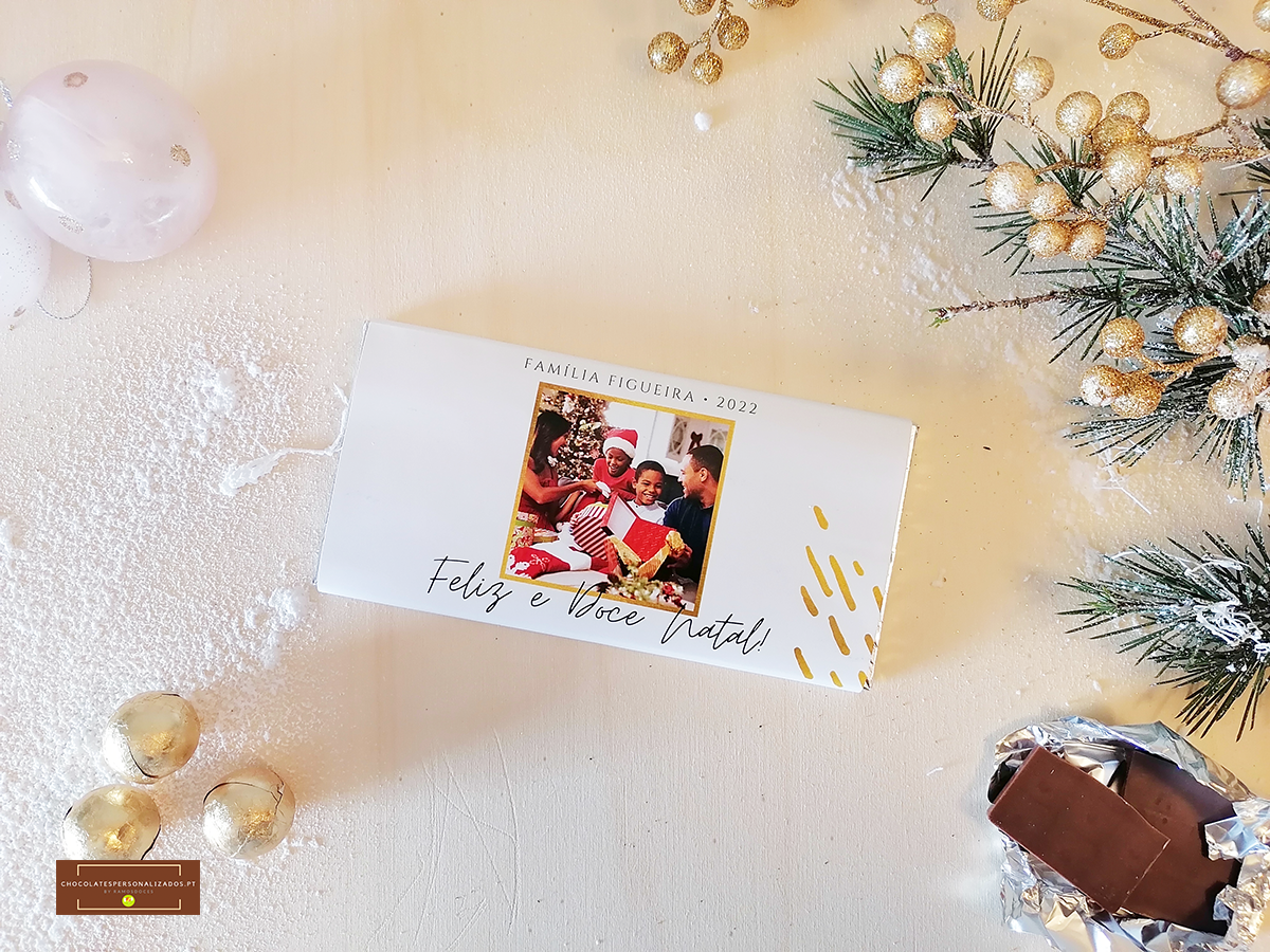 Embalagem branca de chocolate com foto de família e texto de Natal, chocolate parcialmente desembrulhado, decoração natalícia dourada e ramos de pinheiro