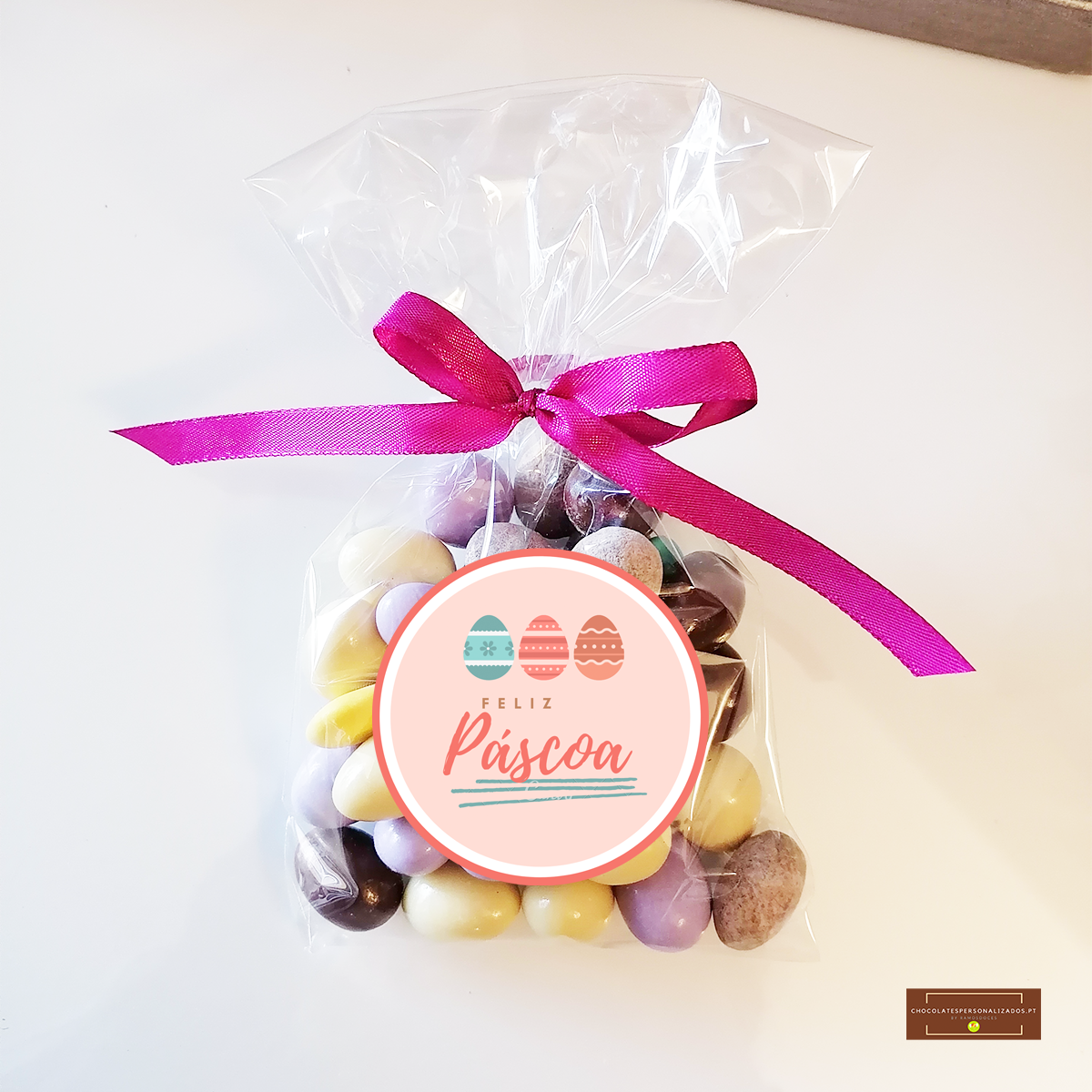 Saco transparente com amêndoas coloridas e laço rosa com etiqueta FELIZ Páscoa