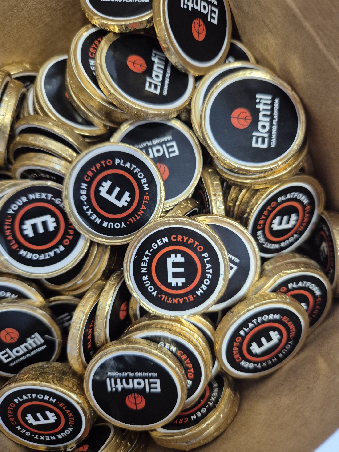 Moedas metálicas douradas com emblemas pretos contendo texto sobre plataforma de criptomoedas dentro de recipiente de papel castanho