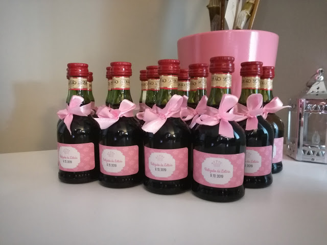 Oito mini garrafas de vidro escuro com rótulo e tampa vermelha e laços cor-de-rosa alinhadas numa superfície branca, com vaso rosa e pincéis ao fundo