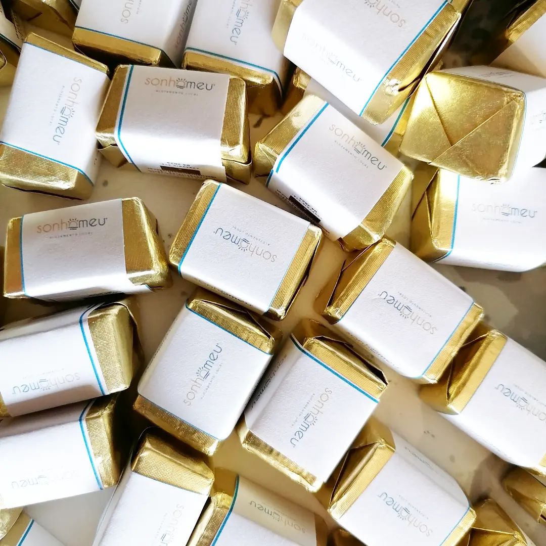 Pequenas embalagens de chocolates quadrados em papel dourado com rótulos brancos com texto 'sonthemeu'