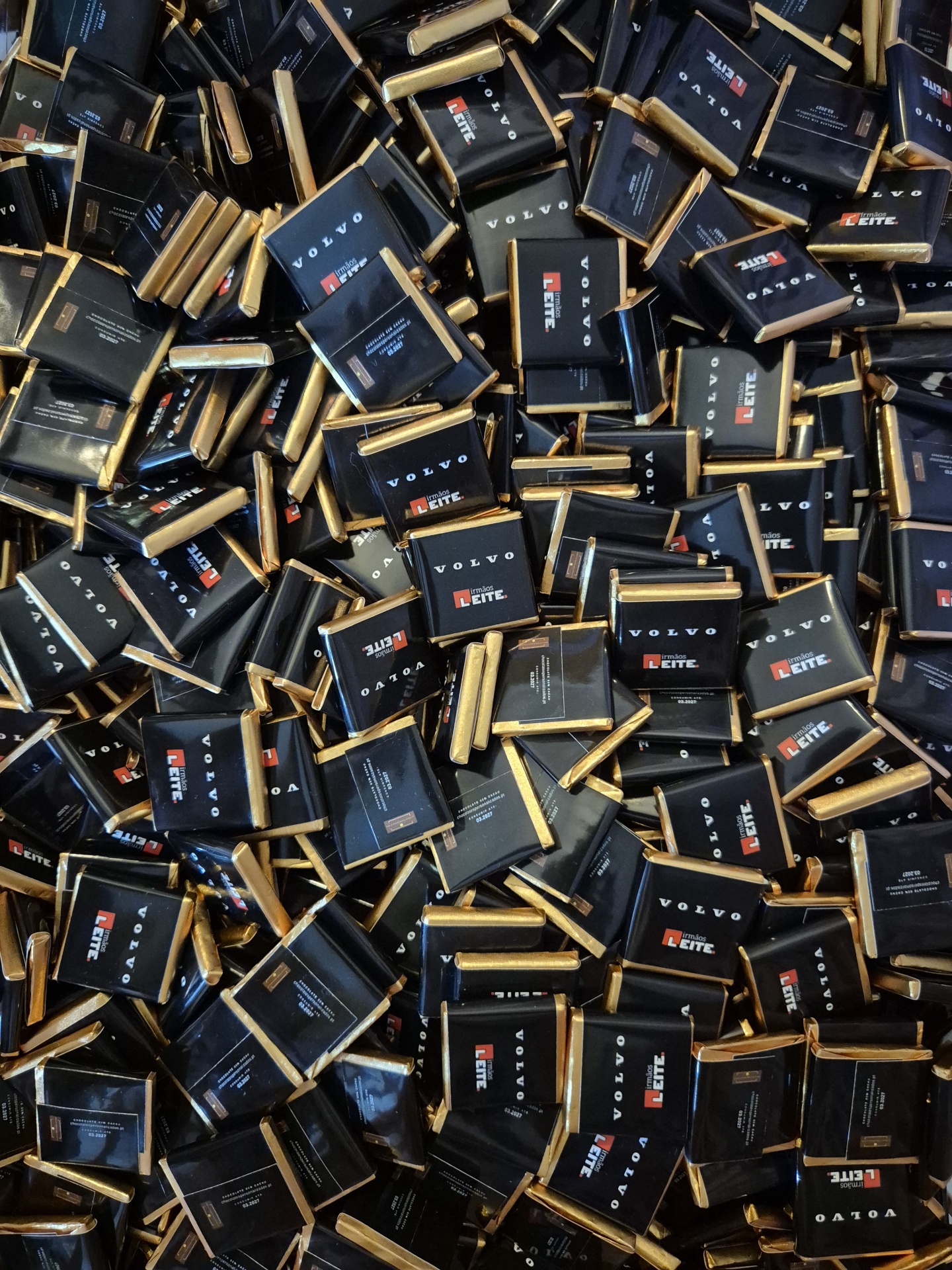Multidão de pequenos chocolates embalados em papel preto com detalhes dourados, texto VOLVO e irmãos LEITE.