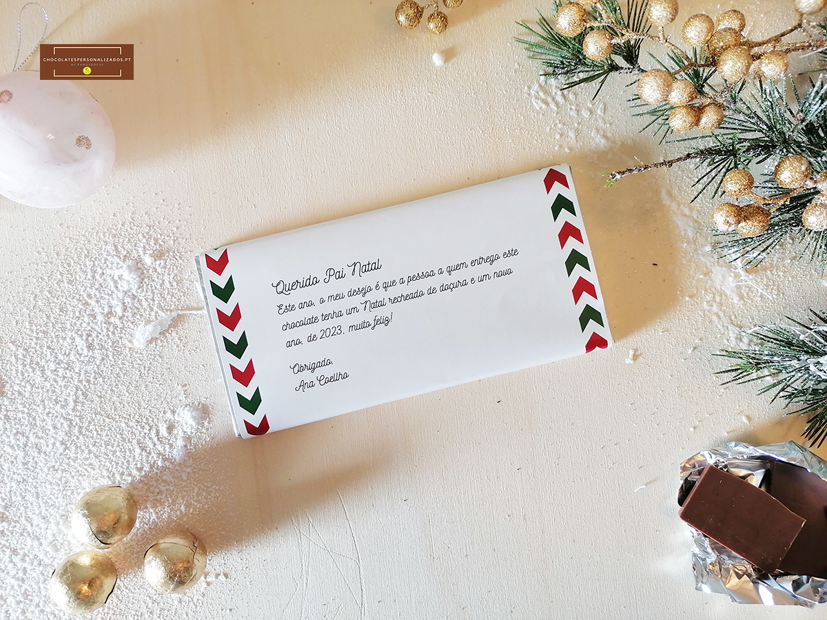 Barra de chocolate com embalagem branca e detalhes vermelhos e verdes, texto em português e decoração natalícia ao fundo