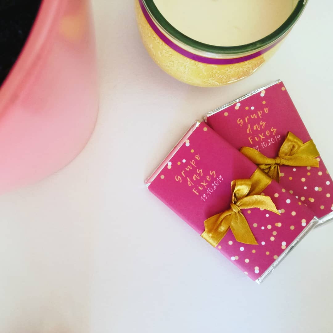 Dois pacotes cor de rosa com laços dourados e texto em amarelo ao lado de vaso rosa e copo decorado