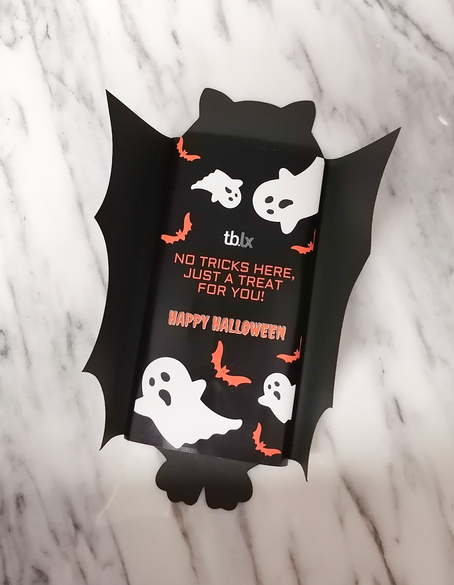 Embalagem preta de Halloween com fantasmas, morcegos e texto em vermelho sobre fundo de mármore