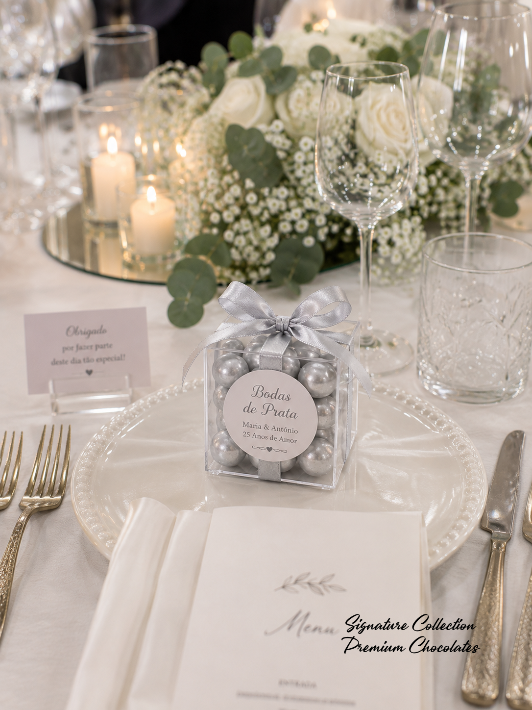 Caixa transparente com bolas prateadas e laço em cima de prato branco numa mesa decorada para celebração.