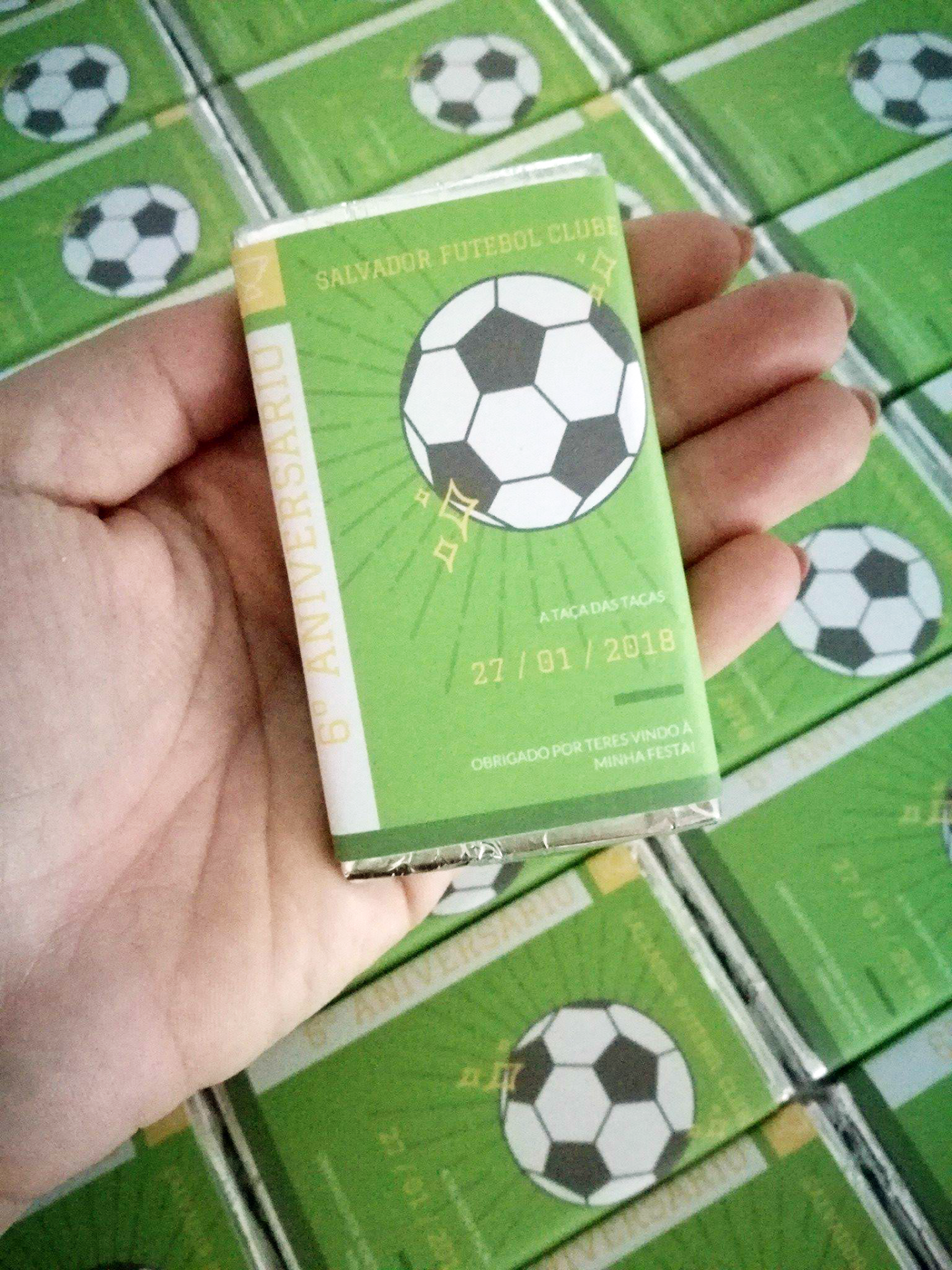 Caixa pequena verde com bola de futebol e texto em português, em mão sobre várias caixas iguais