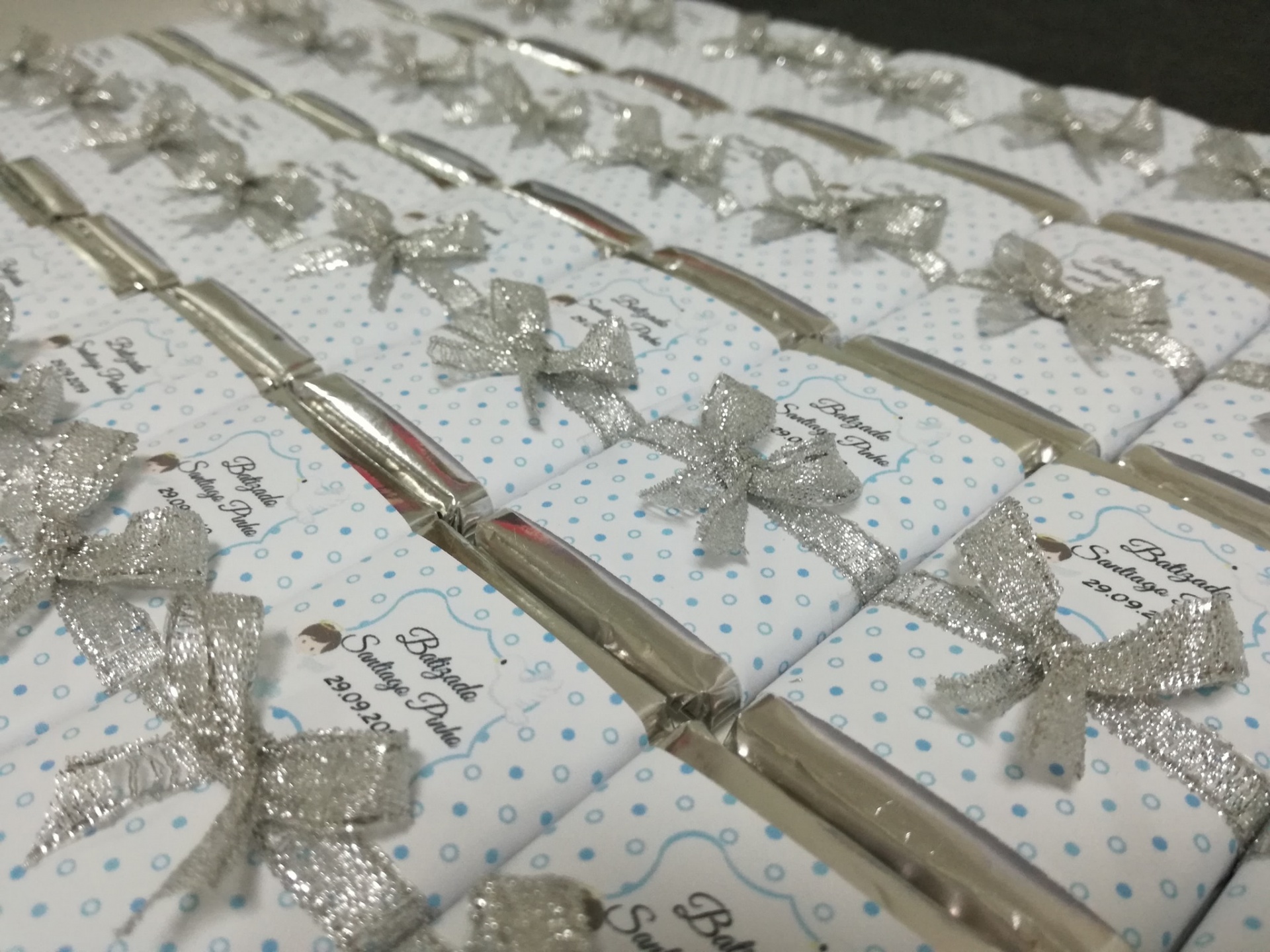 Barras de chocolate embrulhadas com papel branco e bolinhas azuis, laços prateados e etiquetas para batismo