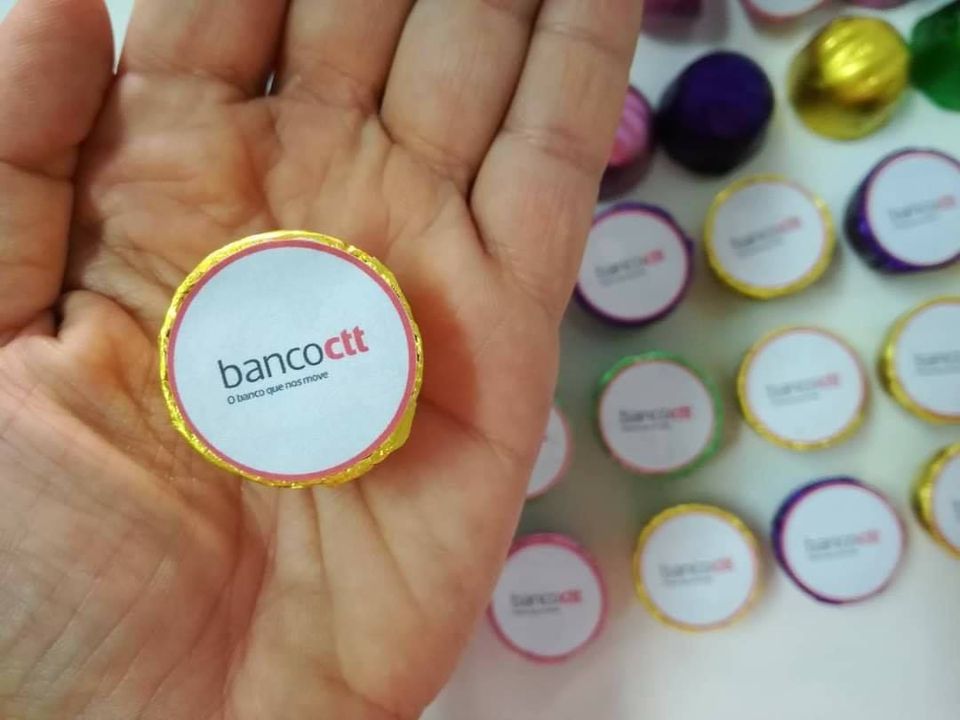 Chocolate redondo com rótulo banco ctt segurado numa mão com vários chocolates semelhantes ao fundo