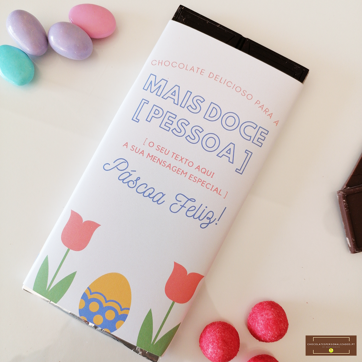 Barra de chocolate com embalagem branca decorada para Páscoa, com texto personalizado e imagens de tulipas e ovo decorado, rodeada por ovos de chocolate coloridos e rebuçados rosas.