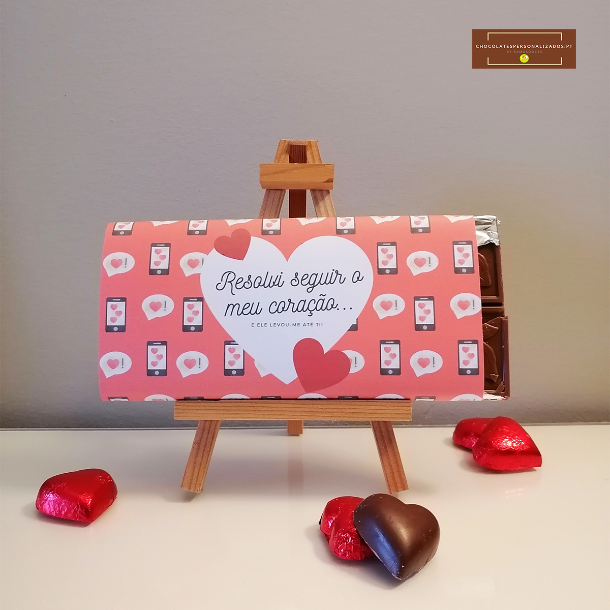 Barra de chocolate em embalagem rosa com corações e texto, em cavalete com bombons de coração vermelhos