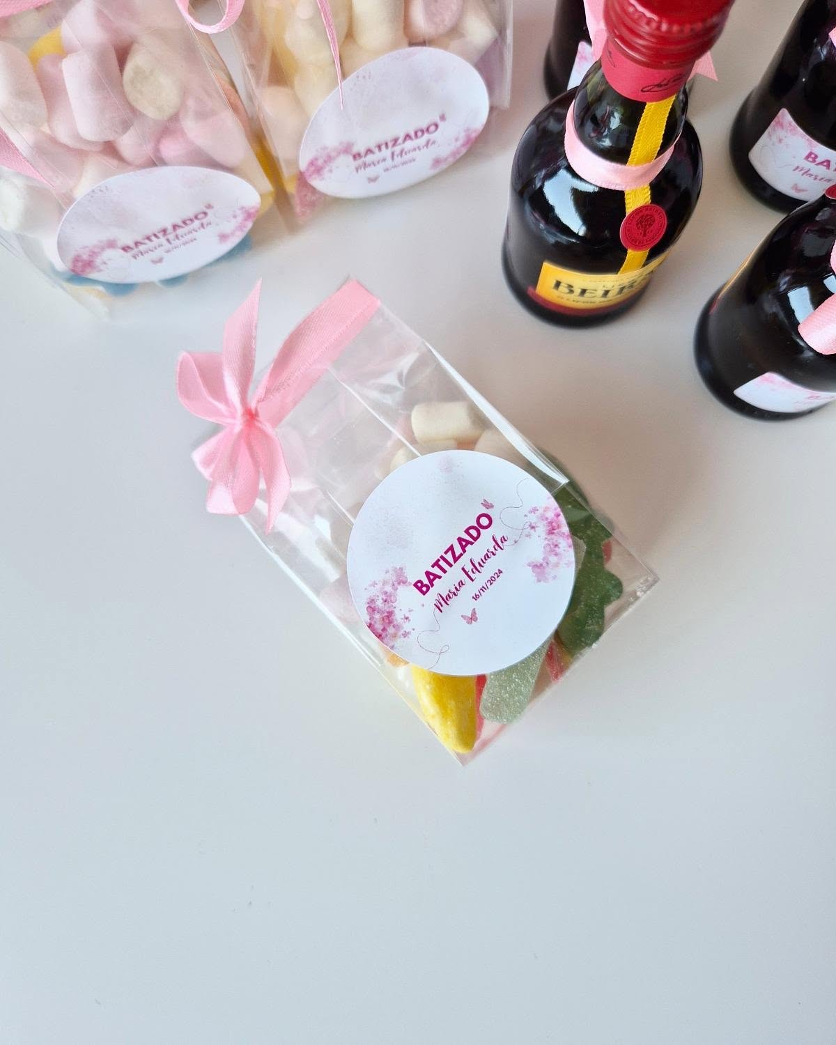 Embalagens de doces coloridos com etiqueta BATIZADO Pink Garden e fita rosa, ao lado de pequenas garrafas de bebida com tampa vermelha
