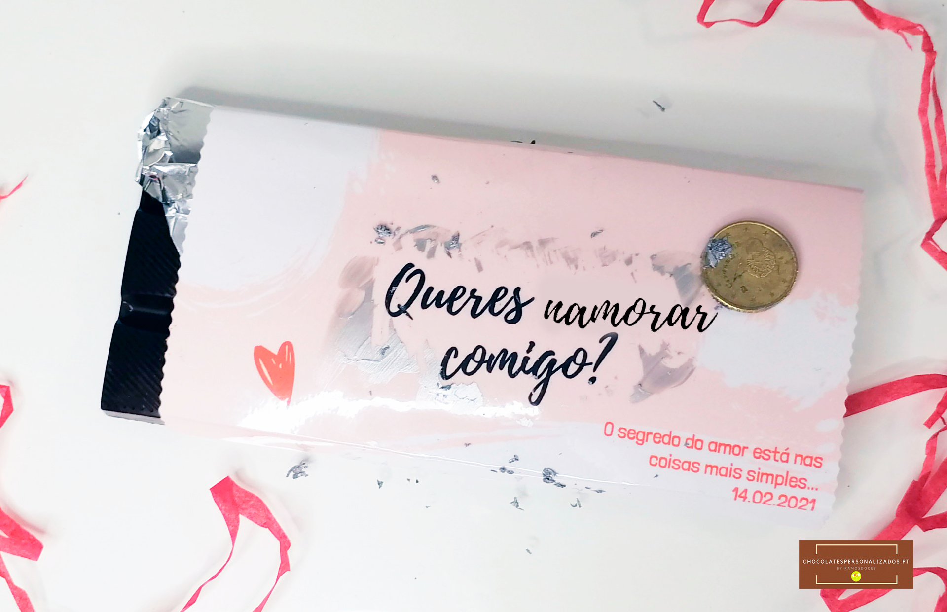 Barra de chocolate com embalagem rosa claro e texto romântico, moeda sobre a barra, fundo branco com fitas vermelhas.