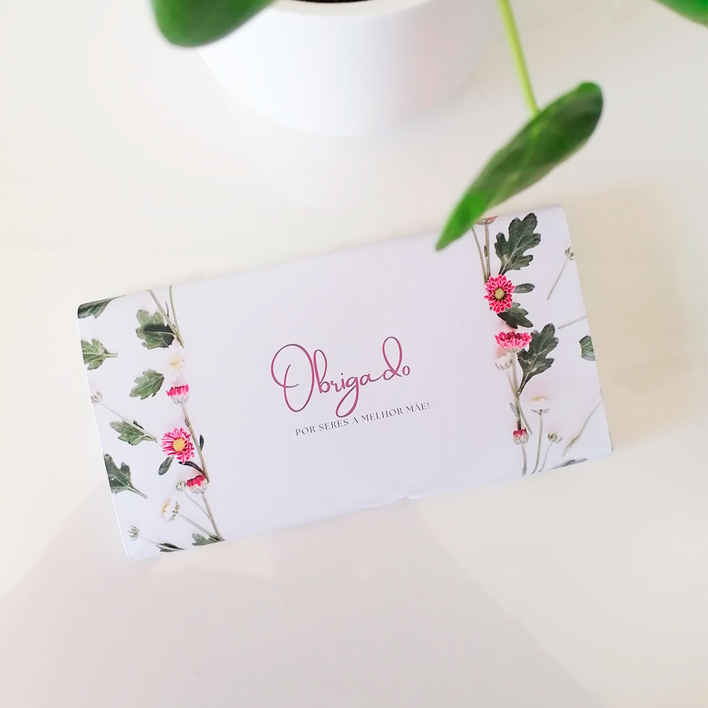 Embalagem branca com folhas e flores cor-de-rosa, texto de agradecimento em português, sobre superfície branca com planta ao fundo