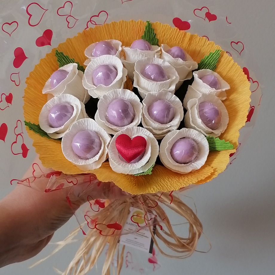 Buquê de cápsulas lilás com decoração em papel amarelo e laço de ráfia, fundo com corações vermelhos.