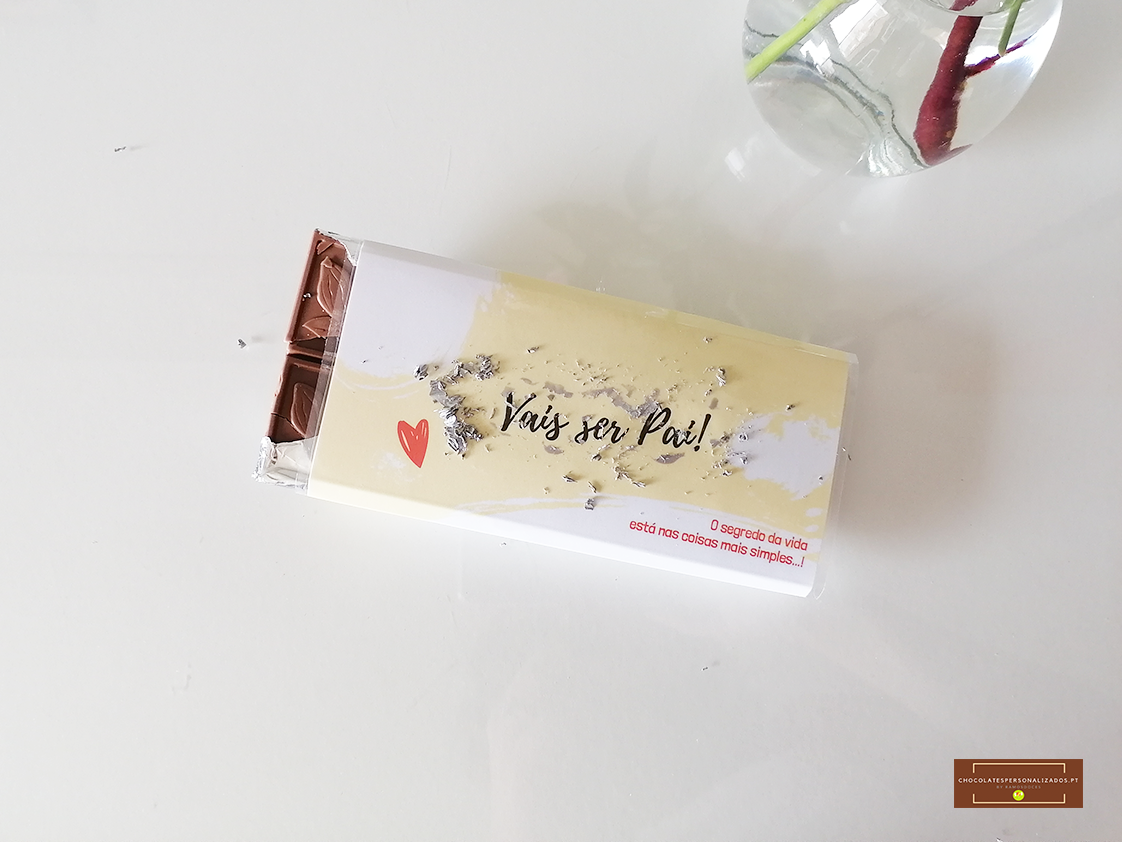 Barra de chocolate com embalagem 'vais ser Pai!' ao lado de um vaso de vidro com flor