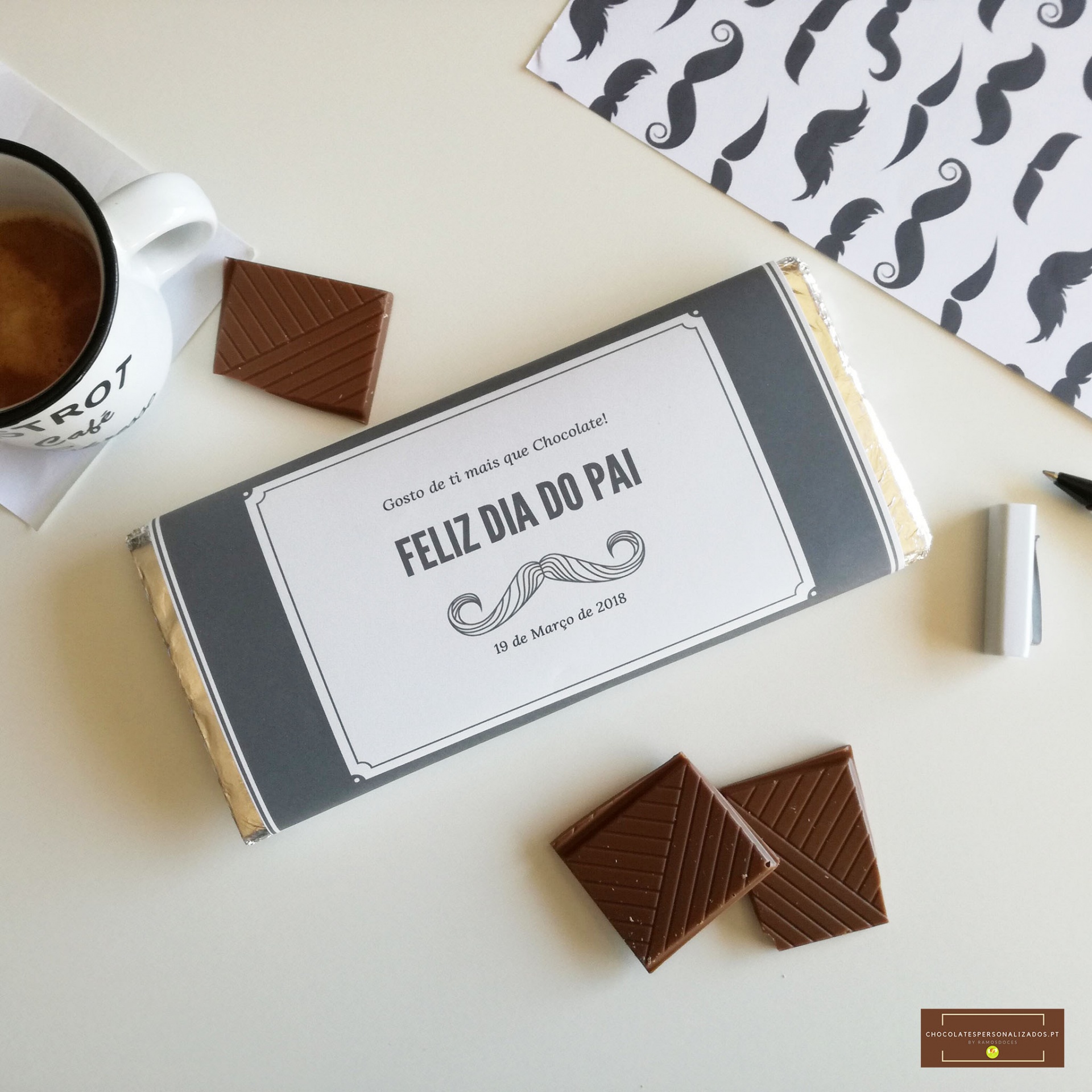 Barra de chocolate com embalagem cinza e branca e texto Feliz Dia do Pai, com pedaços de chocolate e uma chávena de café sobre uma mesa branca
