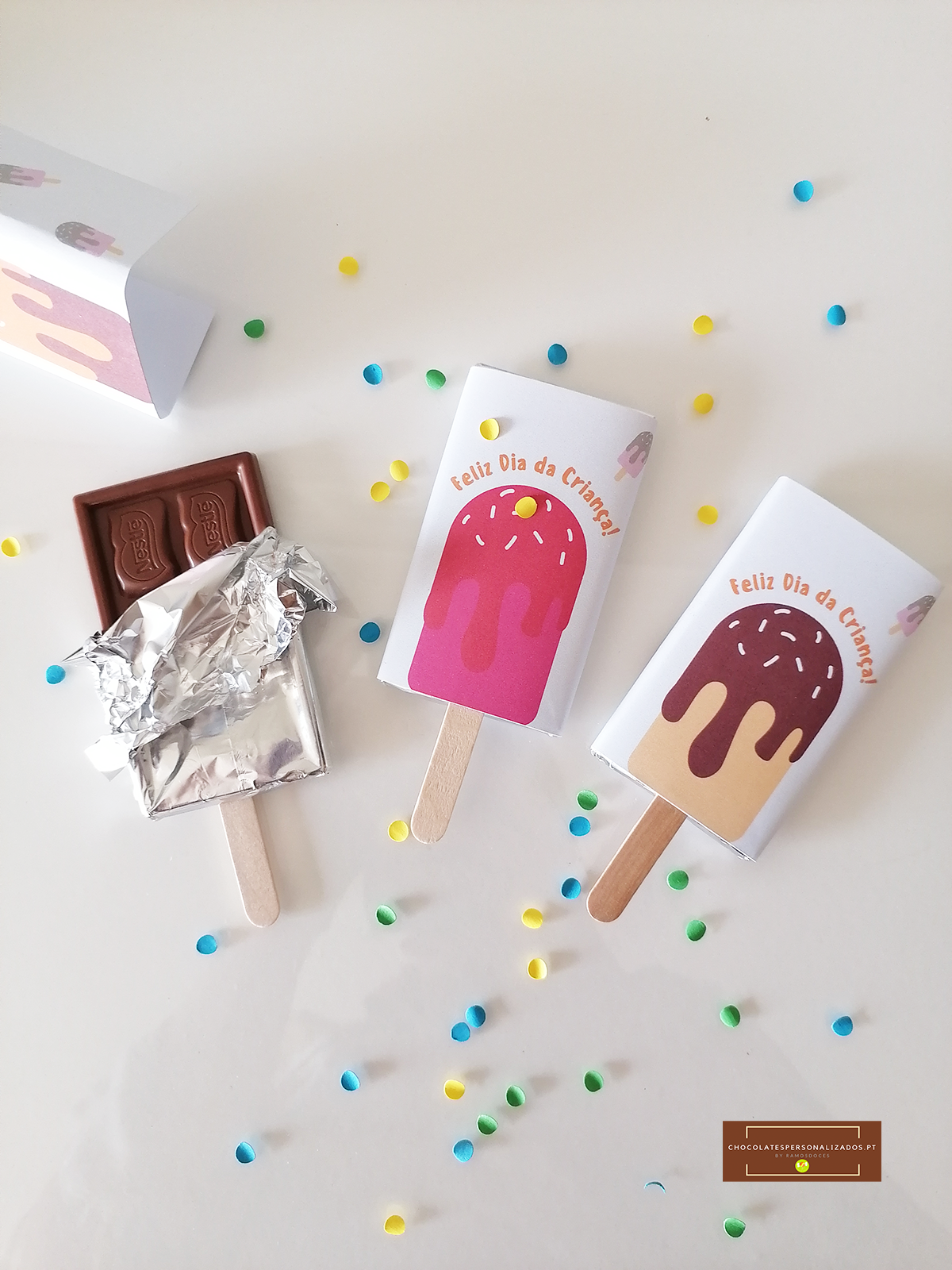 Três chocolates em forma de gelado com embalagem decorativa e confetes coloridos espalhados sobre fundo branco.