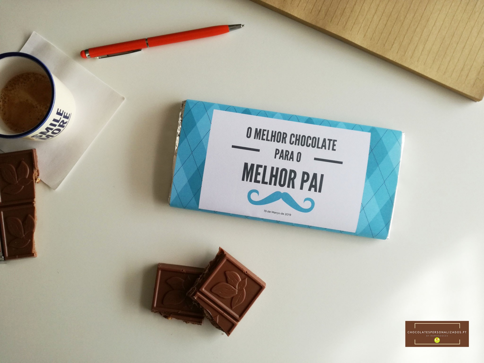 Barra de chocolate embrulhada com texto para o melhor pai, chocolate partido, caneta, caderno e chávena com bebida quente
