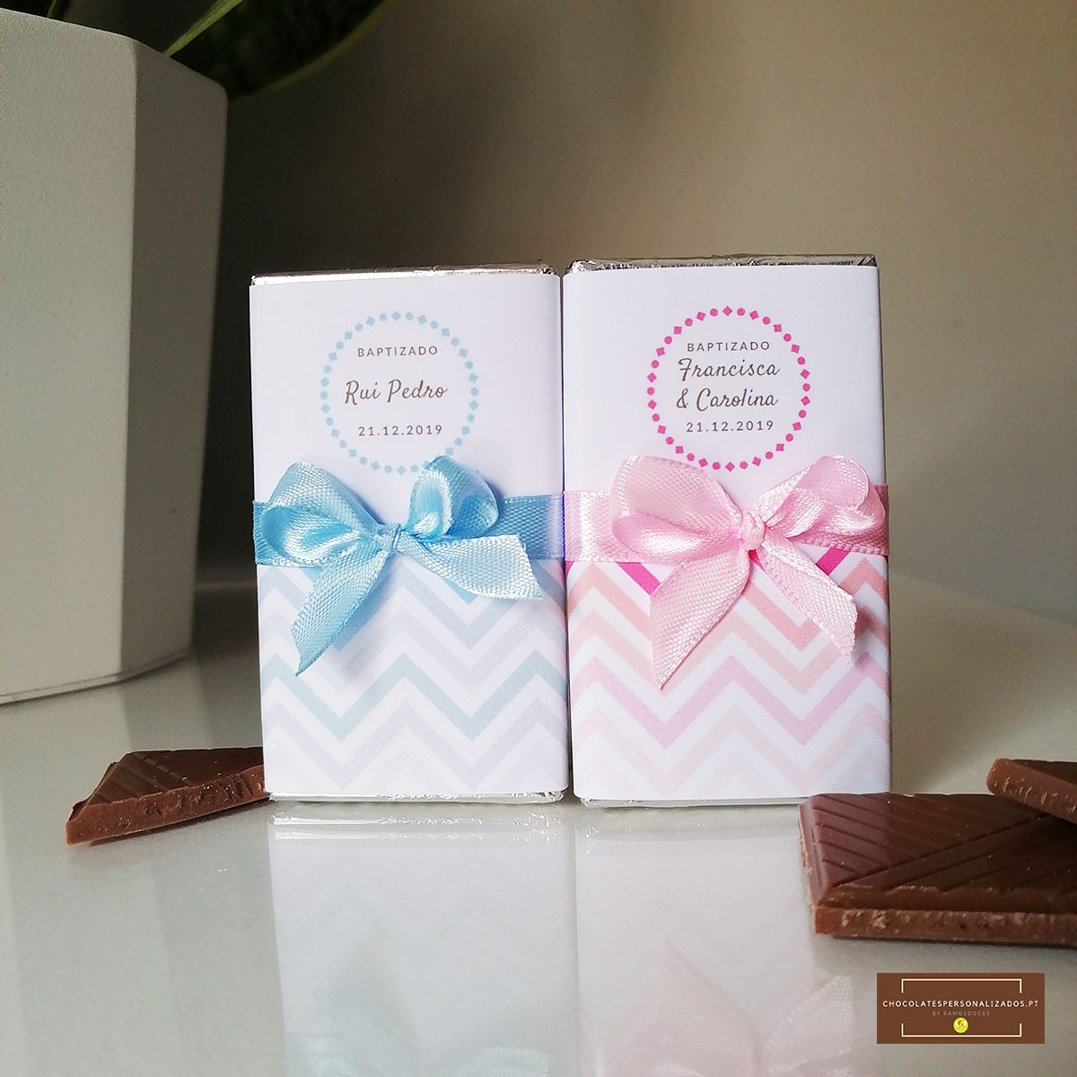 Duas caixas de chocolate embrulhadas com laços azul e rosa sobre superfície branca com pedaços de chocolate.