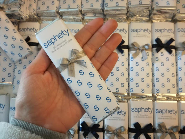 Caixa branca com padrão azul, laço cinza e texto 'saphety' segurada por mão, várias caixas iguais ao fundo