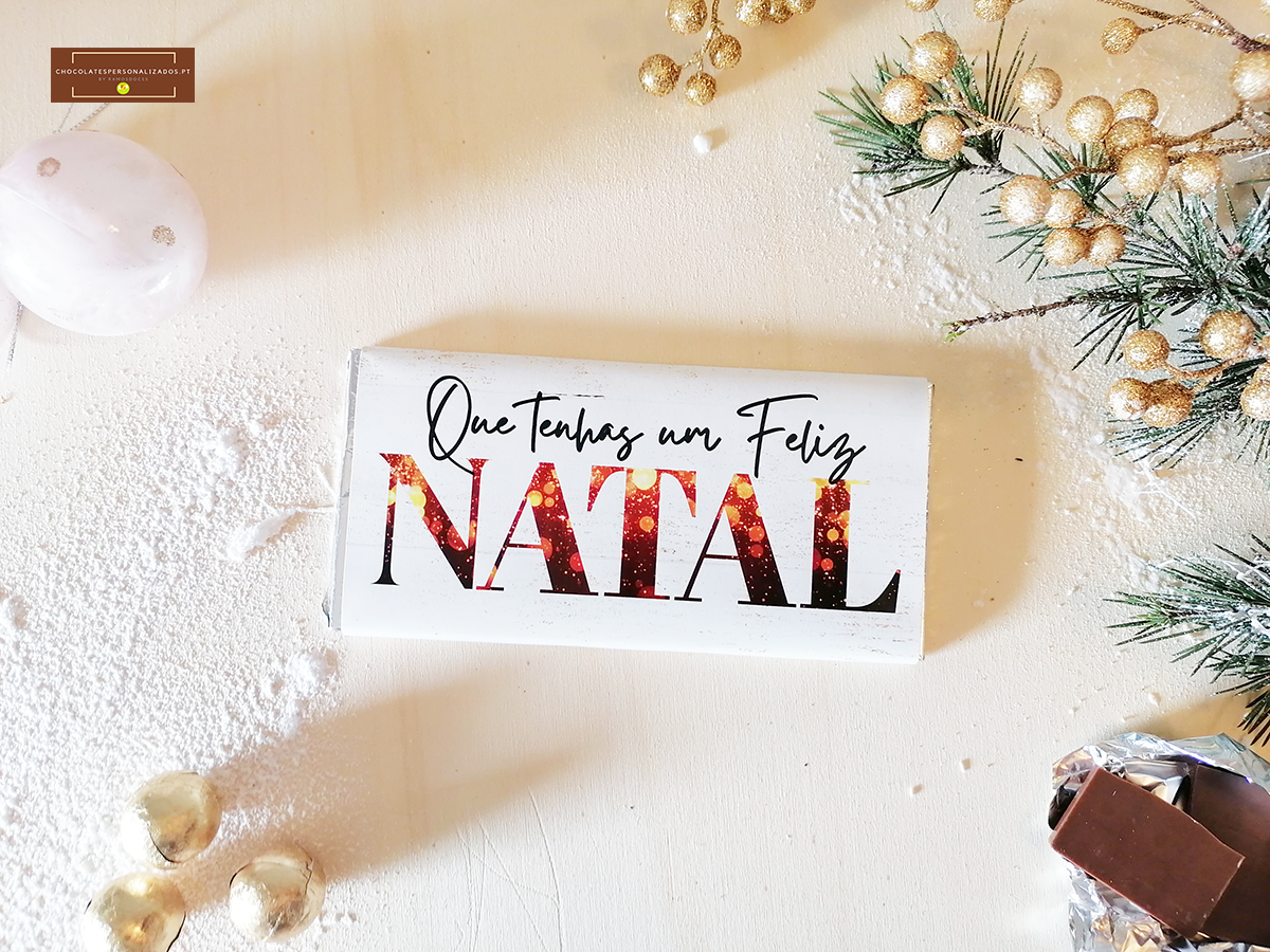 Barra de chocolate com embalagem branca e texto natalício, decoração dourada e galhos verdes com neve artificial.