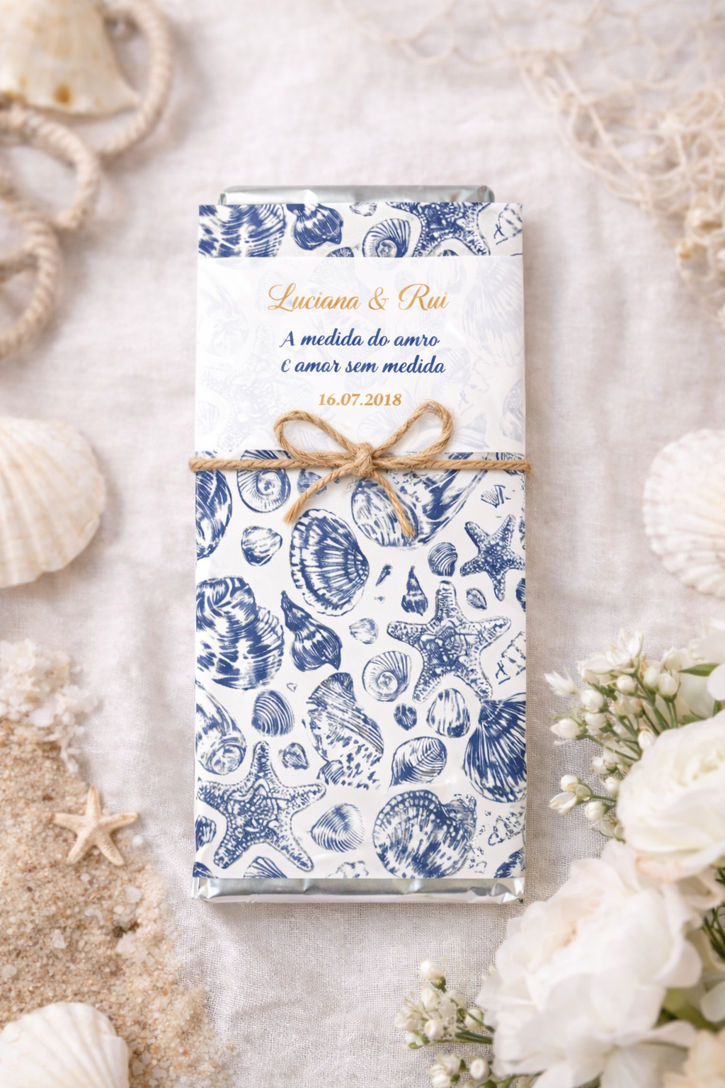 Chocolate embrulhado em papel decorativo com conchas azuis e fio de juta com flores e areia ao redor