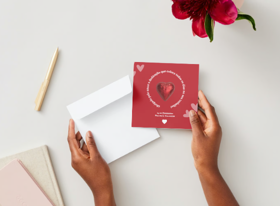 Cartão vermelho com coração brilhante e texto branco em mesa branca com envelope, caneta, cadernos e flor vermelha