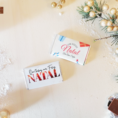 Dois chocolates embrulhados com mensagens de Natal sobre mesa decorada com neve artificial e ramos de pinheiro dourados.