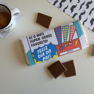 Barra de chocolate com embalagem colorida e texto de Dia do Pai ao lado de chávena de café e pedaços de chocolate