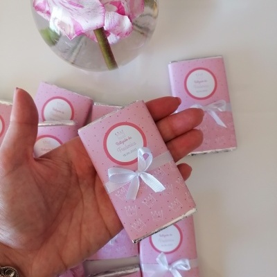 Pequenos chocolates embrulhados em papel rosa com etiquetas e fitas brancas numa mão e sobre uma mesa branca