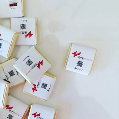 Pequenos chocolates com papel branco, etiqueta com logotipo vermelho, QR code e texto sobre fundo branco.