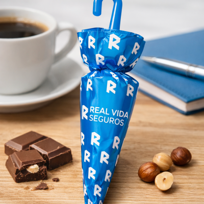 Embalagem azul com letras R e texto REAL VIDA SEGUROS junto a chocolate, avelãs, café e caderno numa mesa