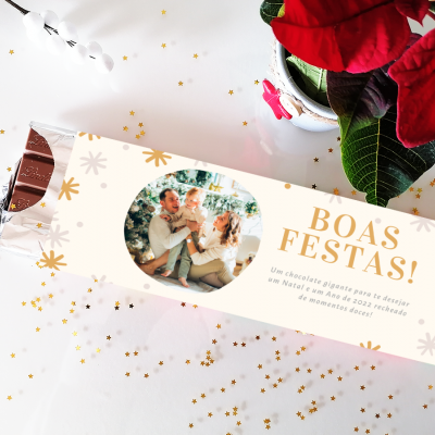 Barra de chocolate com embalagem branca, mensagem Boas Festas e foto de casal, decorada com estrelas douradas, ao lado de planta com flores vermelhas