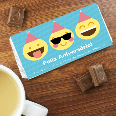 Barra de chocolate com embrulho azul e emojis sorridentes, texto de aniversário, pedaços de chocolate e chávena branca