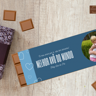 Barra de chocolate com embalagem azul e foto de idoso com criança, ao lado de caixa de chocolate decorada e quadrados de chocolate soltos