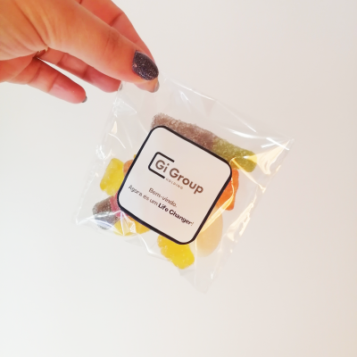 Sacos de Gomas personalizados com logotipo da empresa Mão segurando pacote transparente com balas coloridas e rótulo Gi Group Portugal