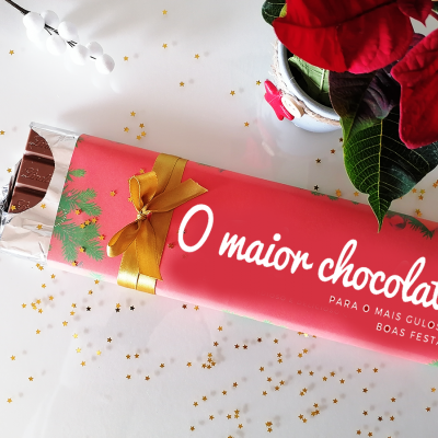 Barra de chocolate com embalagem vermelha e laço dourado, ao lado de planta com folhas vermelhas e estrelas douradas espalhadas
