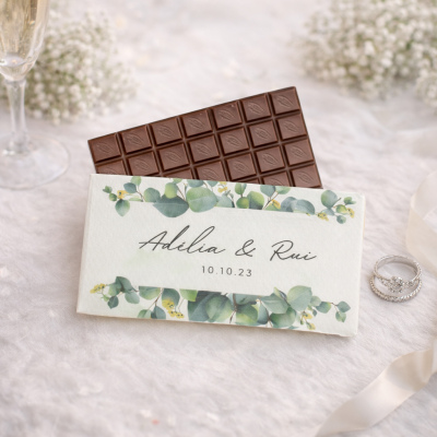Barra de chocolate com embalagem personalizada de folhas verdes e texto Adélia & Rui 10.10.23 ao lado de alianças