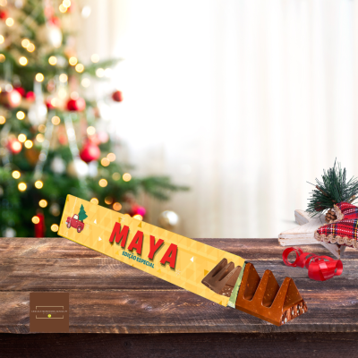 Barra de chocolate Maya em embalagem amarela, sobre mesa de madeira, com decoração natalícia ao fundo.