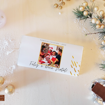 Embalagem branca de chocolate com foto de família e texto de Natal, chocolate parcialmente desembrulhado, decoração natalícia dourada e ramos de pinheiro