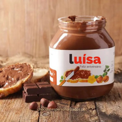 Frasco de creme de chocolate com rótulo branco e texto 'luísa Feliz aniversário' sobre mesa de madeira com chocolate, avelãs e pão
