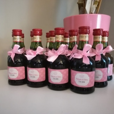 Oito mini garrafas de vidro escuro com rótulo e tampa vermelha e laços cor-de-rosa alinhadas numa superfície branca, com vaso rosa e pincéis ao fundo