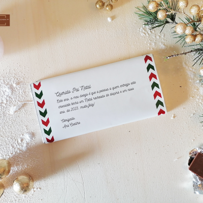 Barra de chocolate com embalagem branca e detalhes vermelhos e verdes, texto em português e decoração natalícia ao fundo